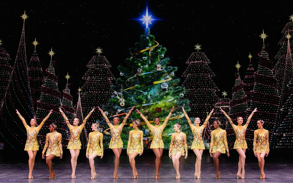 Rockettes in gold costumes performing beside a Christmas tree on the Radio City stage