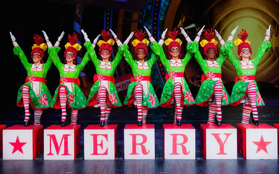 Rockettes in festive costumes beside blocks spelling Merry on stage
