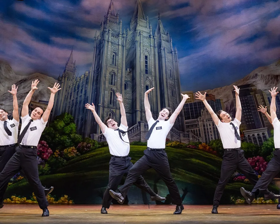 The Book of Mormon ensemble performing before a painted city backdrop
